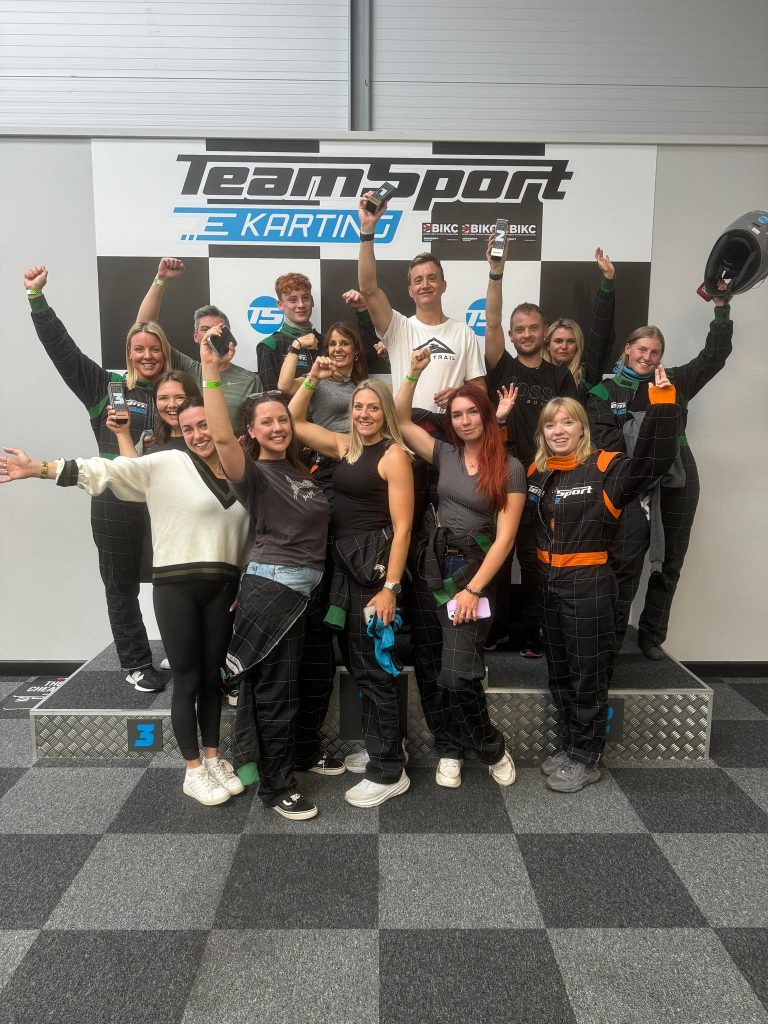 A group of people in racing suits celebrating on a podium at an indoor go-karting venue, standing in front of a "TeamSport Karting" sign, with some raising their arms and one person holding a helmet.