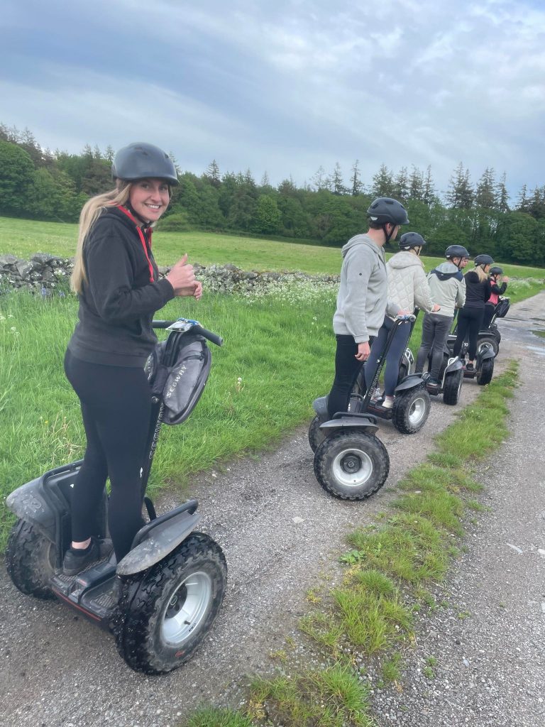 A group of individuals on Segways travelling single file down a rural path, bordered by grass and wildflowers, with a backdrop of green fields and woodland.